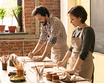 Young creative man and woman in workwear bending over wooden table while working over new collection of earthenware