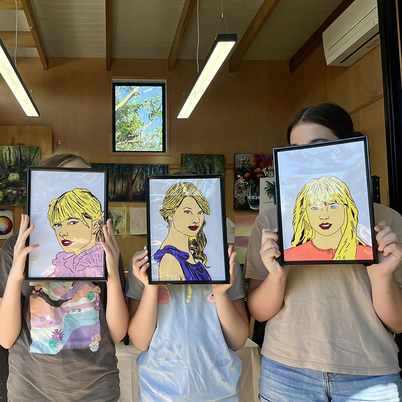 Three girls holding painting of girl portraits in front of their faces.