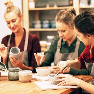 Three cheerful young female friends are painting ceramics in a pottery workshop. Have fun doing art.