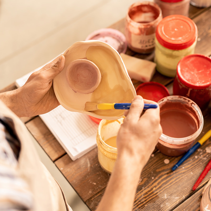 Designer of pottery holding clay workpiece and applying paint from one of plastic jars over workplace