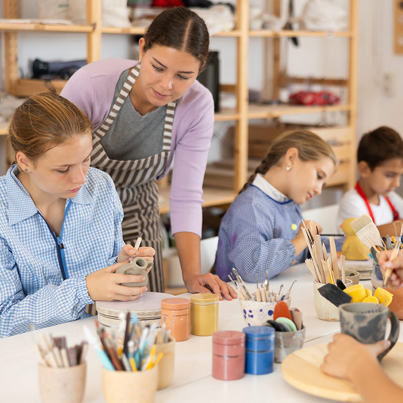 Children and teenagers sit at a table and paint ceramics at the workshop. Teacher shows how to paint a clay product after firing.