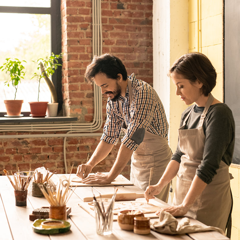 Young creative man and woman in workwear bending over wooden table while working over new collection of earthenware